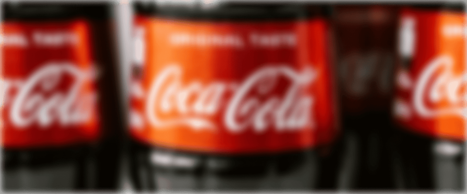 Stark weichgezeichnete Nahaufnahme eines roten Coca-Cola-Etiketts mit dem charakteristischen weißen Markenschriftzug, flankiert von weiteren verschwommenen Flaschen im Hintergrund.