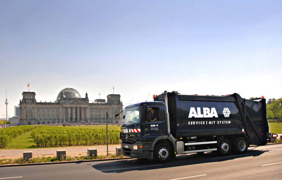 Ein dunkelblauer ALBA-Müllwagen auf einer Straße vor dem historischen Reichstagsgebäude in Berlin unter klarem, blauem Himmel.