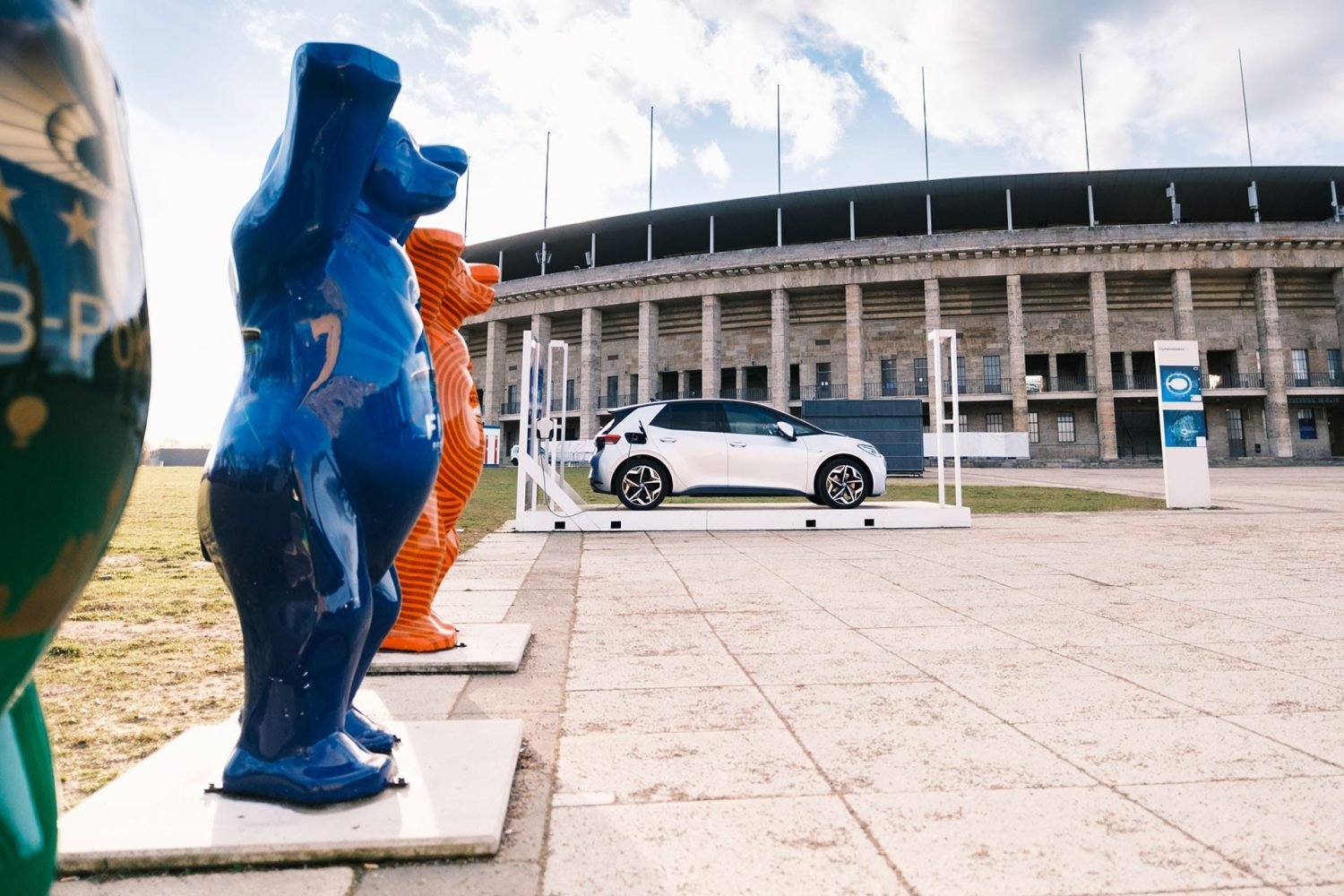 Ein silbernes Elektroauto auf einer Präsentationsbühne vor dem Olympiastadion Berlin, im linken Vordergrund flankiert von den typischen Berliner Buddy-Bär-Skulpturen.