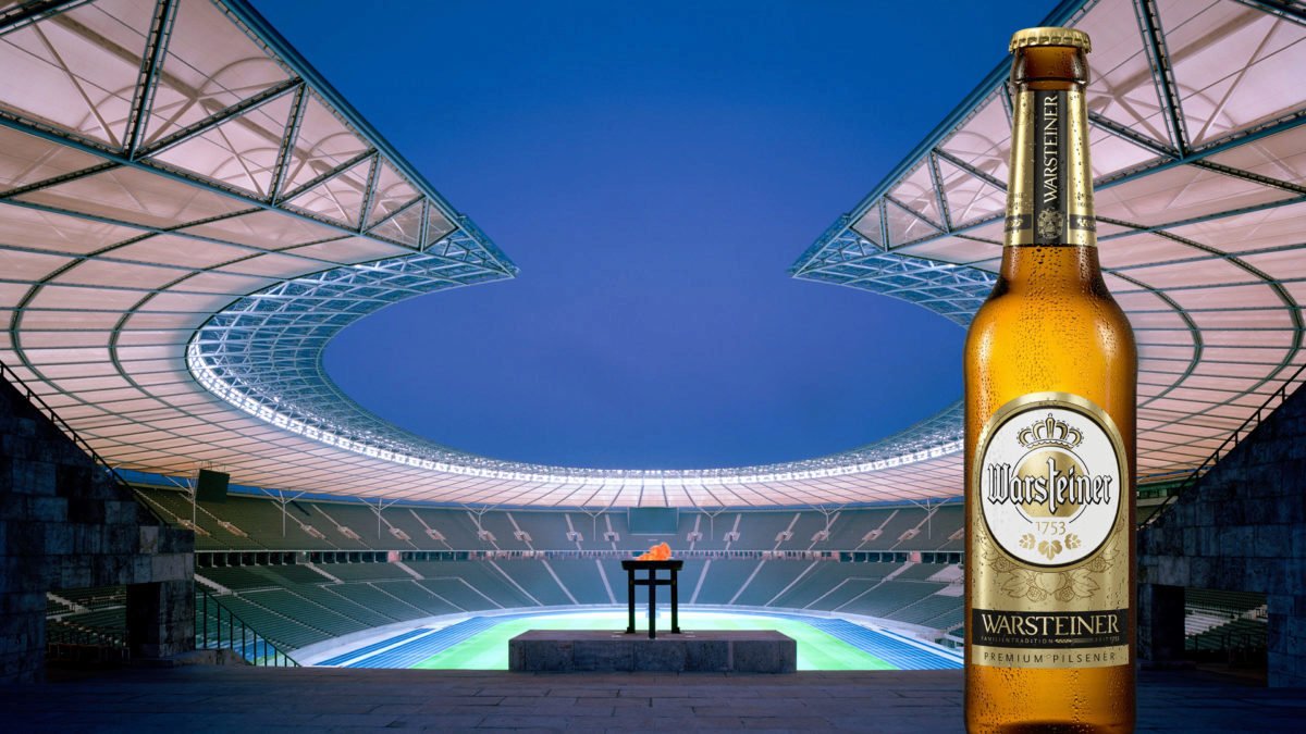 Werbebild für Warsteiner Pilsener: Eine eiskalte, beschlagene Bierflasche steht im Vordergrund vor der abendlichen Kulisse des Olympiastadions Berlin mit brennendem Feuer in der Opferschale am Marathontor.