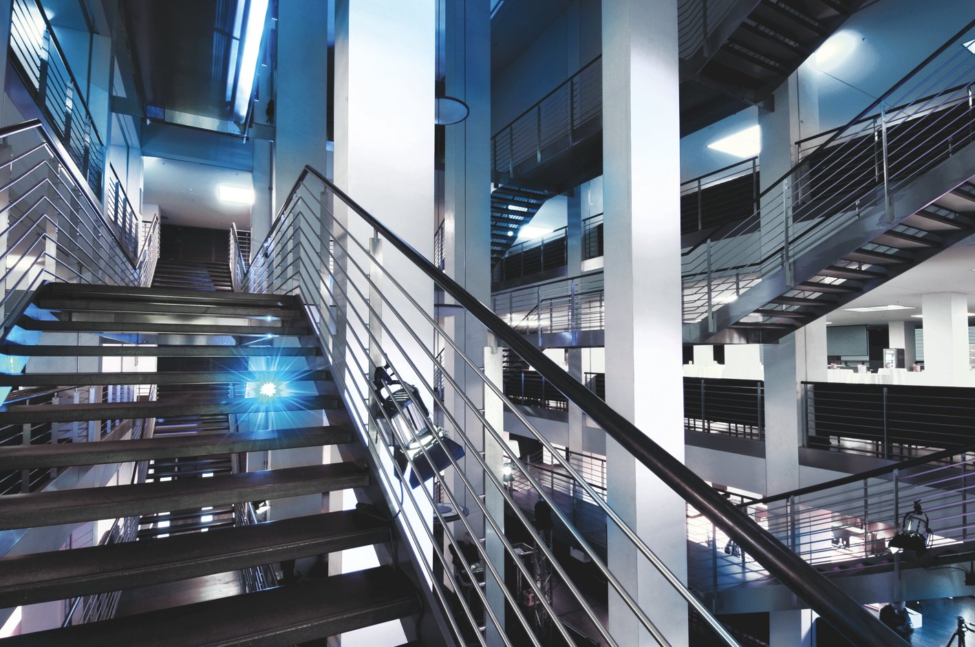 Modernes Atrium im Stadioninnenbereich mit mehreren offenen Treppenfluchten und Geländern aus Edelstahl, dramatisch inszeniert in kaltblauem Licht.
