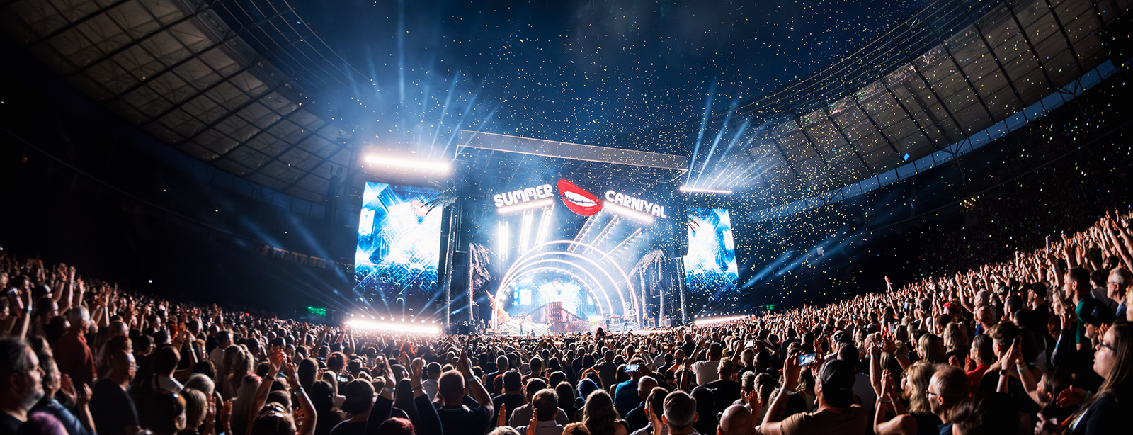 Tausende begeisterte Fans feiern bei einem nächtlichen Stadionkonzert der „Summer Carnival“-Tour im Olympiastadion Berlin vor einer gigantischen, hell illuminierten Showbühne mit spektakulären Lichteffekten und riesigen Leinwänden.