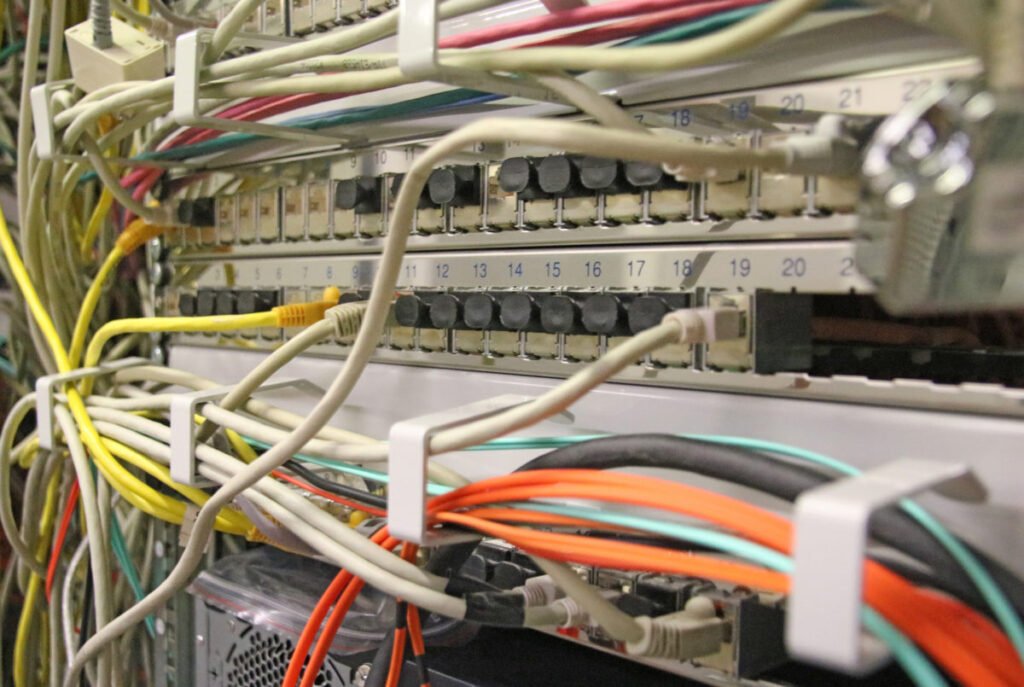 Nahaufnahme eines IT-Netzwerkschranks mit mehreren Patchpanels. Zahlreiche bunte Netzwerkkabel (darunter graue, gelbe, orangefarbene und rote Ethernet-Kabel) verlaufen durch weiße Kabelführungsbügel und sind teilweise in die nummerierten Ports eingesteckt. Nicht belegte Ports sind mit schwarzen Staubschutzabdeckungen versehen. Das dichte Kabelgewirr vermittelt einen Eindruck von komplexer Netzwerkinfrastruktur.
