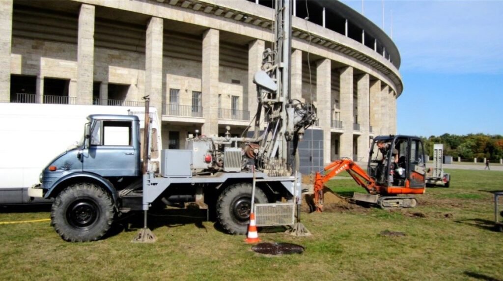 Bohrarbeiten vor der historischen Kulisse des Olympiastadions Berlin mit einer mobilen Brunnen- oder Erdbohranlage auf einem Unimog und einem orangefarbenen Minibagger auf der Rasenfläche.