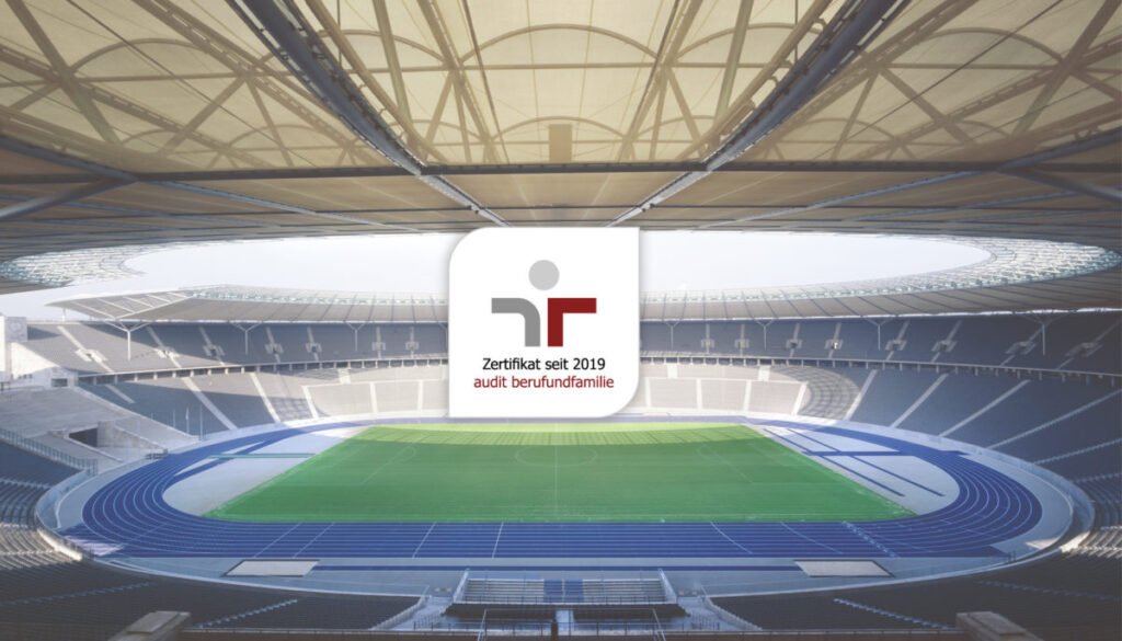 Das Logo des Zertifikats „audit berufundfamilie“ mit dem Zusatz „Zertifikat seit 2019“ liegt als Einblendung über einer Innenansicht des Olympiastadions Berlin mit Rasenfläche und blauer Laufbahn.