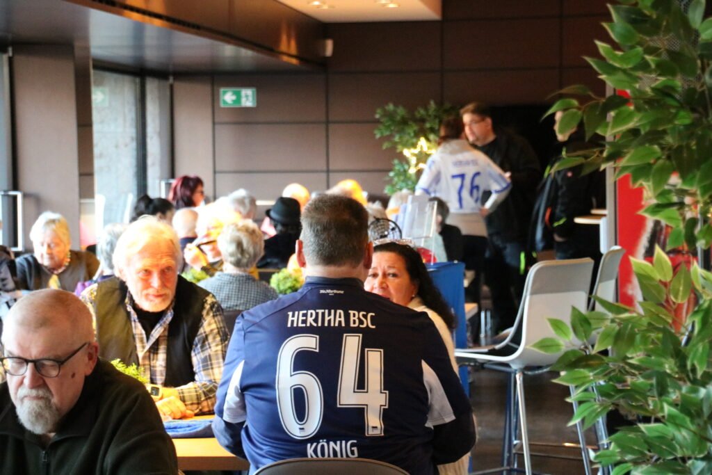 Belebter VIP- oder Loungebereich im Olympiastadion Berlin mit Fans an den Tischen, im Vordergrund ein Hertha BSC Fan im dunkelblauen Trikot mit der Nummer 64.