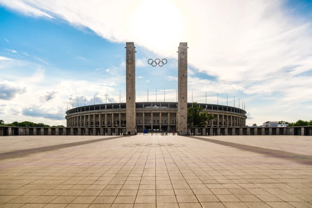 Weitwinkel-Außenaufnahme des Osttors am Olympiastadion Berlin an einem sonnigen Tag. Im Zentrum ragen die beiden monumentalen, freistehenden Steintürme empor, zwischen denen an Stahlseilen die historischen Olympischen Ringe hängen. Ein großer, gepflasterter Vorplatz führt fluchtend auf das Stadiontor zu. Dahinter erstreckt sich die halbrunde, säulenartige Natursteinfassade des Stadions unter einem strahlend blauen Himmel mit weichen, weißen Wolken, während die Sonne direkt hinter den Ringen strahlt.