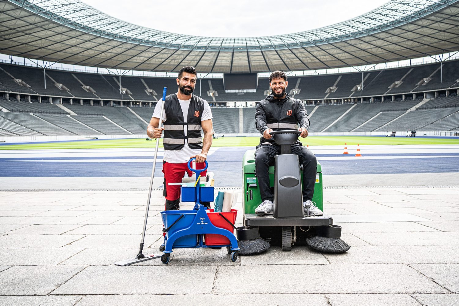 Zwei Reinigungskräfte posieren lächelnd mit einem Putzwagen, einem Mopp und einer grünen Aufsitz-Kehrmaschine im Innenraum des Olympiastadions Berlin.